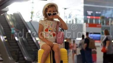 小女孩游客坐在火车站的一个大黄色手提箱上。 儿童正在等待出发，前往